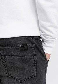 Jeans de mezclilla negra con un acabado texturizado, que cuentan con un bolsillo trasero con un parche de logo de cuero negro y detalles de costura en contraste.