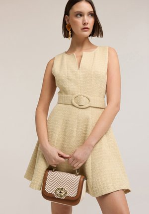 Femme portant une robe sans manches beige texturée avec ceinture, tenant un petit sac à main marron et crème à motif avec une fermeture dorée.