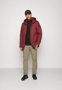 Rode pufferjas met een capuchon, zwart vest met rits en lichtbeige cargobroek. Zwarte schoenen en een pet met witte tekst maken de look compleet.