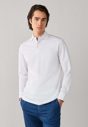 Homme portant un polo blanc à manches longues et un pantalon bleu, debout avec les mains dans les poches devant un fond uni.