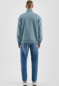 Felpa azzurro chiaro con collo alto, abbinata a jeans denim azzurro chiaro e sneakers bianche, vista da dietro.