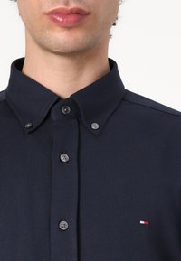 Tummansininen polo-paita, jossa on napillinen kaulus, teksturoitu kangas ja viisi nappia edessä. Sisältää pienen brodeeratun logon punaisella, valkoisella ja sinisellä.