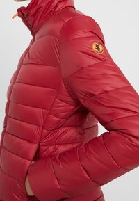 Rode donsjas met horizontale doorgestikte lijnen, hoge kraag en ritssluiting aan de zijkant. Heeft een ronde oranje logo op de linkerschouder.