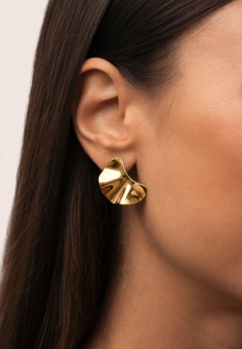 Pendientes de metal dorado con un diseño escultórico y ondulante, que presentan un acabado de alto brillo y una forma asimétrica, llevados en la oreja de un modelo.