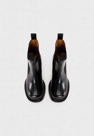 Une paire de bottes Chelsea en cuir noir avec des languettes de tirage et des semelles épaisses, présentée de dessus sur un fond clair.