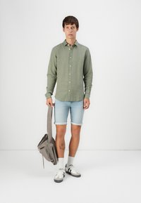 Camisa verde de manga larga abotonada, pantalones cortos de denim azul claro remangados, bolso gris de hombro, calcetines blancos y zapatillas con acentos negros.