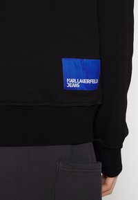 Sudadera negra con un acabado texturizado, que presenta una etiqueta azul con "KARL LAGERFELD JEANS" en letras blancas en la parte inferior.