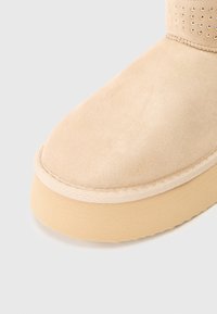 Béžové semišové kotníkové boty s kulatou špičkou, texturovaným povrchem a platformovou podrážkou s jemným šitím a malými studovými akcenty.