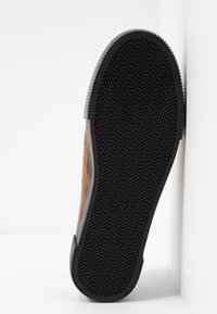Semelle de chaussure avec une semelle extérieure en caoutchouc noir présentant un motif en chevrons, inclinée contre une surface d’angle blanche.
