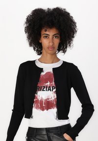 Schwarze, kurz geschnittene Strickjacke mit rundem Ausschnitt und Reißverschluss, getragen über einem weißen T-Shirt mit rotem Grafikdruck und schwarzem Text.