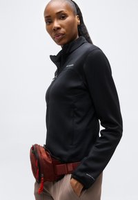 Γυναίκα που φοράει μαύρο μπουφάν με φερμουάρ και μπεζ παντελόνι, στέκεται με το ένα χέρι στην τσέπη και μια κόκκινη ζώνη-τσάντα γύρω από τη μέση.