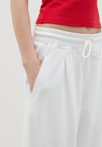 Pantalones de chándal blancos, con plisados, y cintura elástica con cordón. Textura suave, bolsillos laterales, combinados con una camiseta roja ajustada.