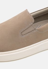 Chaussure beige sans lacets en daim lisse, dotée de panneaux élastiques sur les côtés, d'une semelle blanche et de surpiqûres contrastantes le long des bords.
