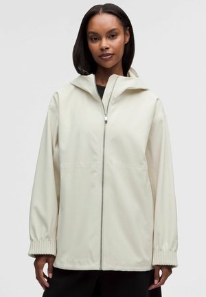 Frau trägt eine hellbeige Jacke mit Reißverschluss, elastischen Bündchen und Kapuze und steht vor einem einfarbigen hellgrauen Hintergrund.