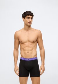 Homem de peito nu com cabelo escuro curto, vestindo boxers pretos com uma faixa roxa da Tommy Hilfiger, de pé contra um fundo branco simples.