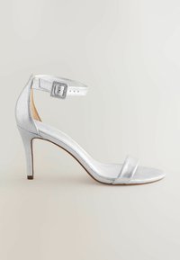 Sølvmetallic højhælede sandaler med ankelrem, en flad tårem og en slank stilettohælf. Glat tekstur og minimalistisk design.