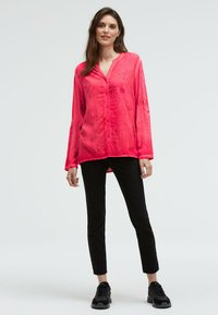 Frau steht mit einer leuchtend rosa Button-Up-Bluse mit dezenten Textmustern, schwarzen engen Hosen und schwarzen Sneakers vor einem weißen Hintergrund.