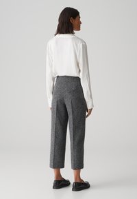 Witte blouse met lange mouwen; hoog aangesloten, grijze textuur broek met een brede pijp. Zwarte instappers. Effen achtergrond.