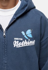 Navy zip-up hoodie made of cotton with a white "Good for Nothing" text and a blue butterfly graphic on the left chest area.