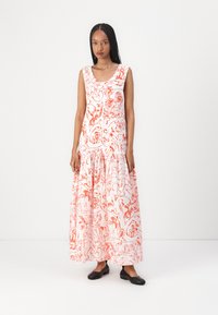 Maxikleid aus weißem Stoff mit roten abstrakten Mustern, ärmelloses Design, Rundhalsausschnitt und gestuftem Saum. Kombiniert mit schwarzen flachen Schuhen.
