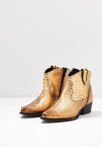 Ankle-high golden boots with a textured, scale-like pattern, pointed toes, side zippers, and stacked black heels on a white surface.