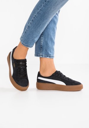 Stopy w czarno-białych butach sportowych Puma z brązowymi podeszwami, zestawione z niebieskimi podwiniętymi dżinsami na białym tle.