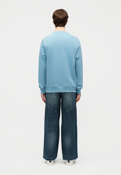 Άτομο που φοράει ανοιζομπλέ πουκάμισο με μακριά μανίκια και σκούρα μπλε τζιν παντελόνι φαρδιάς γραμμής, κοιτάζοντας προς τα πίσω με φόντο έναν απλό λευκό τοίχο.