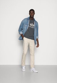 Eine Jeansjacke, ein graues T-Shirt mit Logo, beigefarbene Slim-Fit-Hosen und weiße Sneaker. Das Model steht lässig mit einer Hand in der Tasche.