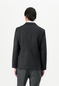 Mann in einem dunklen Hahnentrittmuster-Blazer und grauen Hosen, der mit dem Rücken zur Kamera vor einem schlichten weißen Hintergrund steht.