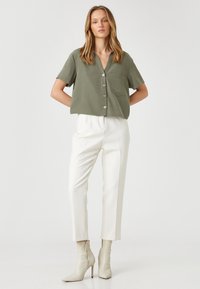 Camisa verde de manga corta con botones y bolsillo, combinada con pantalones blancos ajustados y botas de tobillo puntiagudas en un tono beige claro.