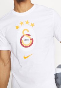 T-shirt bianca con logo Galatasaray giallo e rosso, testo "1905", cinque stelle gialle sopra, baffo Nike giallo sotto, persona che tiene un pallone da calcio.