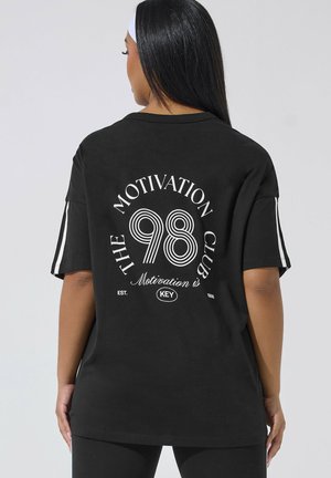 Kobieta o długich czarnych włosach, ubrana w czarny, oversize'owy t-shirt z białymi paskami na rękawach i napisem "The Motivation Club 98" na plecach.