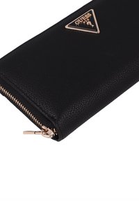 Black leather wallet with a textured finish, gold-toned zipper, and prominent triangular Guess logo accent on the front.