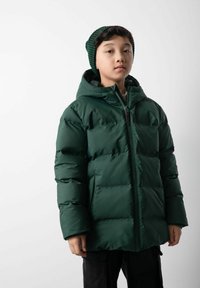 Ciemnozielona kurtka puchowa z zamkiem z przodu i kapturem, z pikowanym wzorem i kieszeniami bocznymi. Noszona z zieloną czapką w paski.