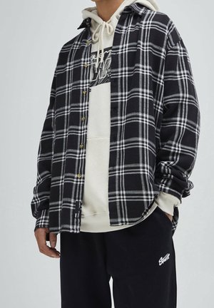 Homme portant une chemise à carreaux noir et blanc par-dessus un sweat à capuche beige avec un pantalon noir avec un texte brodé blanc.