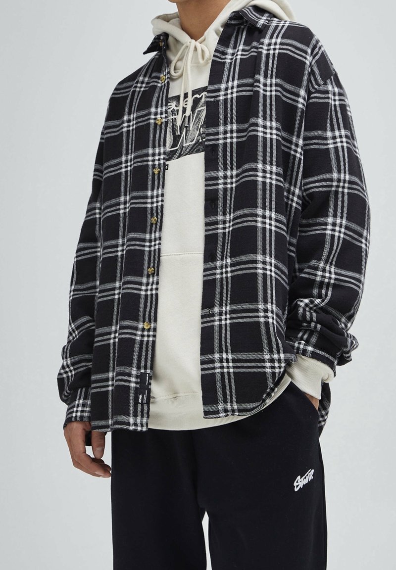 Homme portant une chemise à carreaux noir et blanc par-dessus un sweat à capuche beige avec un pantalon noir avec un texte brodé blanc.