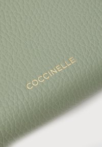 Roheline tekstitud nahkrahakott, mille esiküljel on embosseeritud kuldne "Coccinelle" logo. Sile viimistlus ja mustriline pind.