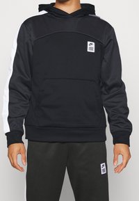 Sweat à capuche noir avec des bandes blanches sur les côtés, une poche kangourou et un patch carré avec le logo Nike sur la poitrine. Matière douce et texturée.