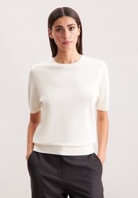 Weißes, gestricktes Kurzarmoberteil mit rundem Ausschnitt, weicher Textur und taillierter Saum, kombiniert mit dunklen Hosen, die einen Kontrast in den Farben schaffen.