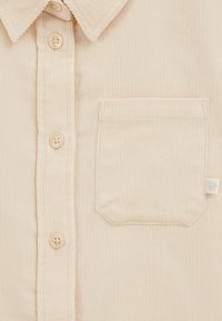 Camicia in corduroy beige con colletto a punta, chiusura frontale con bottoni, taschino singolo sul petto e un piccolo dettaglio a forma di cuore sulla tasca.