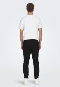 Camiseta blanca de algodón con cuello redondo, combinada con pantalones negros y zapatillas blancas, vista desde la parte trasera sobre un fondo neutro.