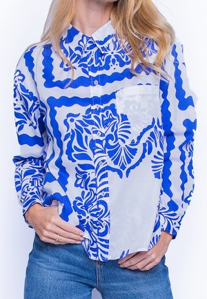 Femme portant une chemise à manches longues boutonnée avec col, à motifs bleus et blancs, rentrée de manière décontractée dans un jean bleu, les mains près de la taille.