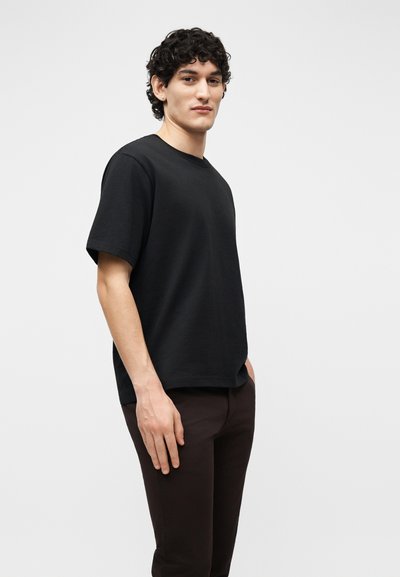 Jeune homme aux cheveux bouclés portant un t-shirt noir à manches courtes et un pantalon sombre, debout devant un fond clair uni.