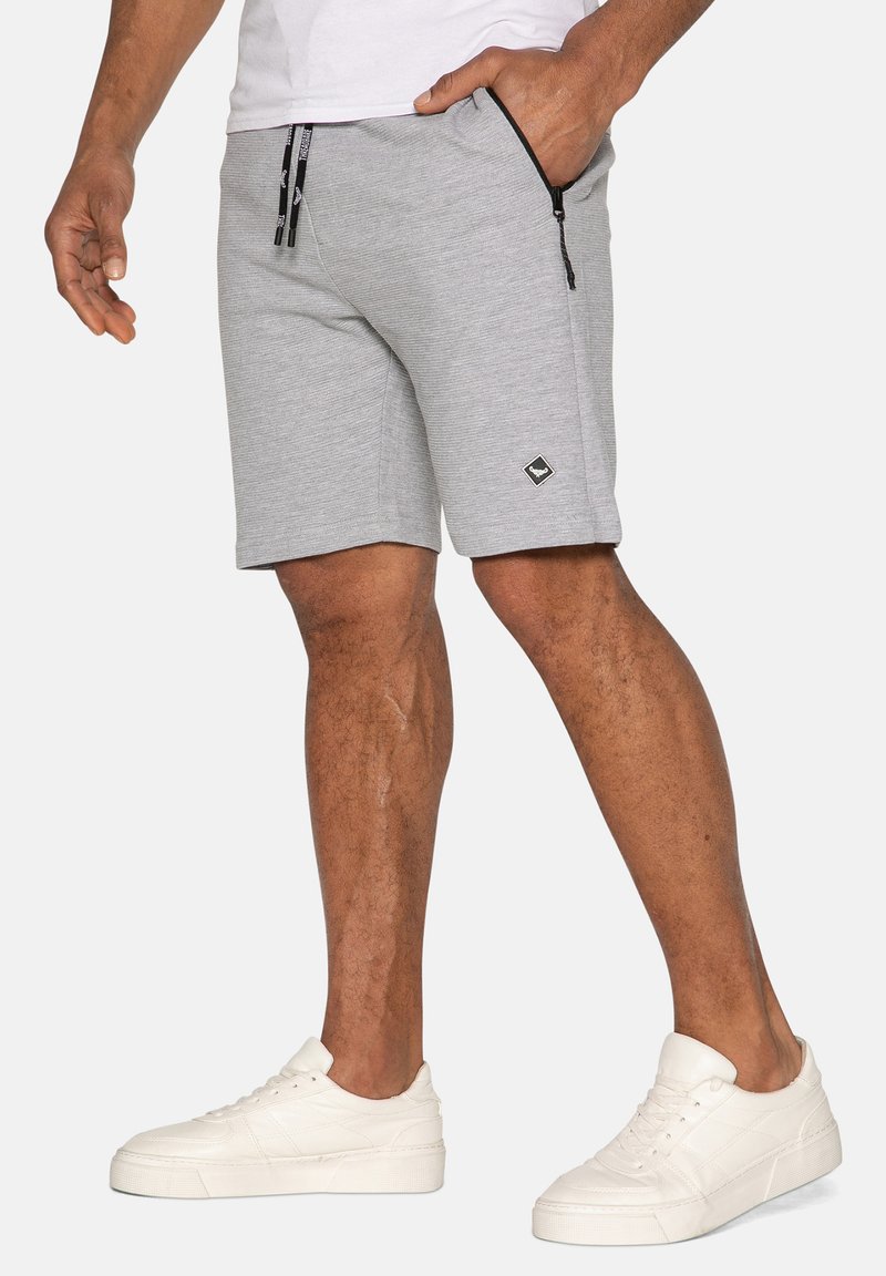 Grå atletiske shorts med en glat tekstur, udstyret med en sort logo-patch, lynlåslommer og en snoreliv. Sammen med hvide sneakers.