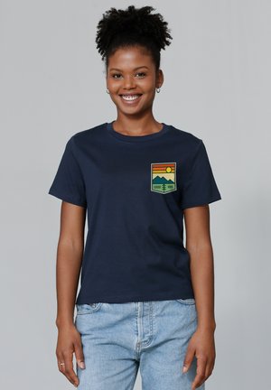 T-shirt en coton bleu marine avec un emblème graphique représentant des montagnes, un soleil et le texte "GOOD VIBE" en vert, jaune et orange.