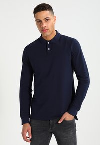 Mørkeblå langermet polo med krage, tre knapper og en jevn tekstur. Den passer tydelig sammen med mørke jeans.