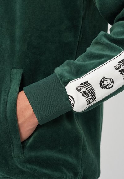 Casaco de veludo verde com acabamento texturizado, punhos canelados e impressão de logotipo ao longo da manga em branco e preto. Bolso lateral visível.