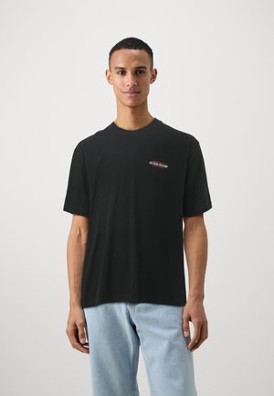 Melnas kokvilnas T-krekls ar īsām piedurknēm, kurā ir neliels sarkans "GUESS JEANS" logo kreisajā krūtī. Vienkārša, brīvā piegriezuma dizains.
