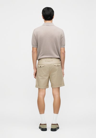Μπεζ βαμβακερό πόλο πουκάμισο συνδυασμένο με χακί σορτς με γυρισμένες ραφές και πίσω τσέπες. Το μοντέλο φοράει λευκές κάλτσες και γκρι παπούτσια.