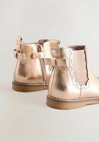 Stivaletti alla caviglia metallici rosa con cerniera e fiocco decorativo. Presentano bordi ondulati e una finitura testurizzata. Suola piatta in gomma per stabilità.